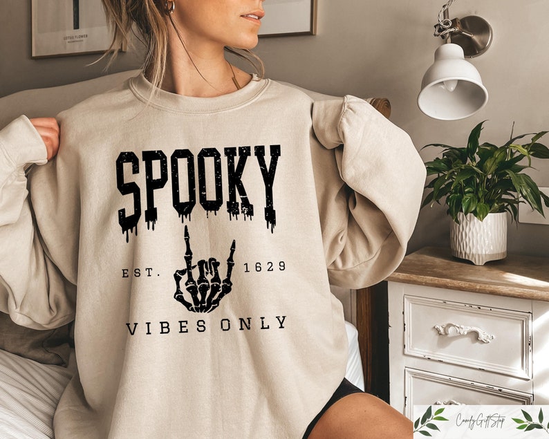 Puede incluir: Una sudadera beige con texto negro que dice "SPOOKY EST. 1629 VIBES ONLY". El texto est&aacute; goteando y una mano de esqueleto est&aacute; haciendo un gesto de rock and roll.