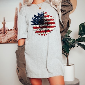 May include: A light gray t-shirt featuring a sunflower design with the American flag colors. The sunflower's petals are red and white stripes, and the center is a dark brown. The shirt is a casual, oversized fit.
