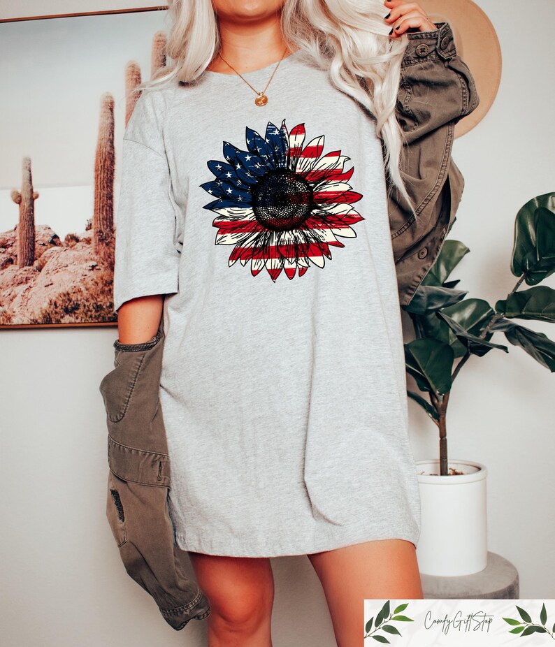 May include: A light gray t-shirt featuring a sunflower design with the American flag colors. The sunflower's petals are red and white stripes, and the center is a dark brown. The shirt is a casual, oversized fit.