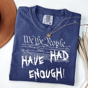 Puede incluir: Una camiseta azul marino Comfort Colors con el texto blanco "We the People HAVE HAD ENOUGH!". Un collar dorado con un colgante descansa sobre la camisa. Un sombrero marr&oacute;n y una hoja de palma seca est&aacute;n en el fondo.