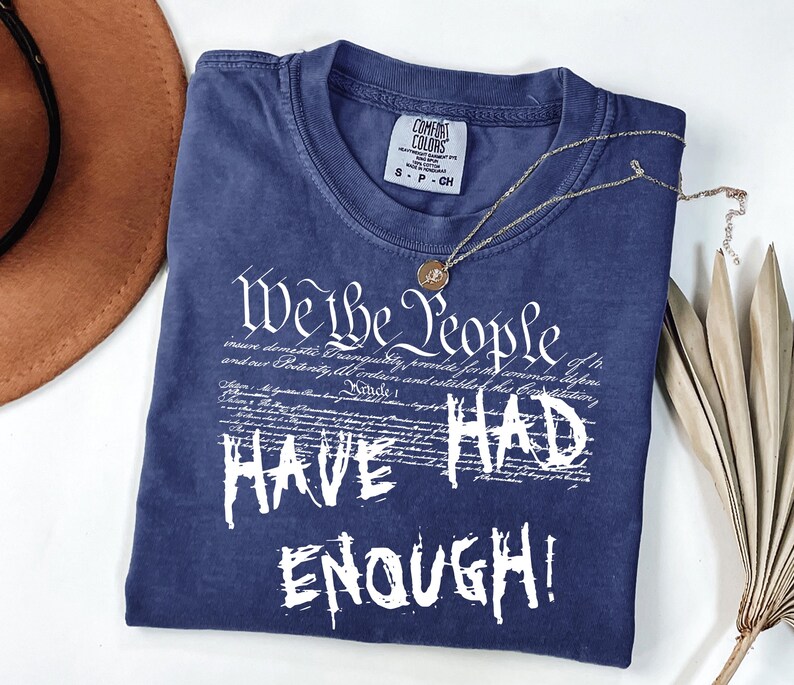Puede incluir: Una camiseta azul marino Comfort Colors con el texto blanco "We the People HAVE HAD ENOUGH!". Un collar dorado con un colgante descansa sobre la camisa. Un sombrero marr&oacute;n y una hoja de palma seca est&aacute;n en el fondo.