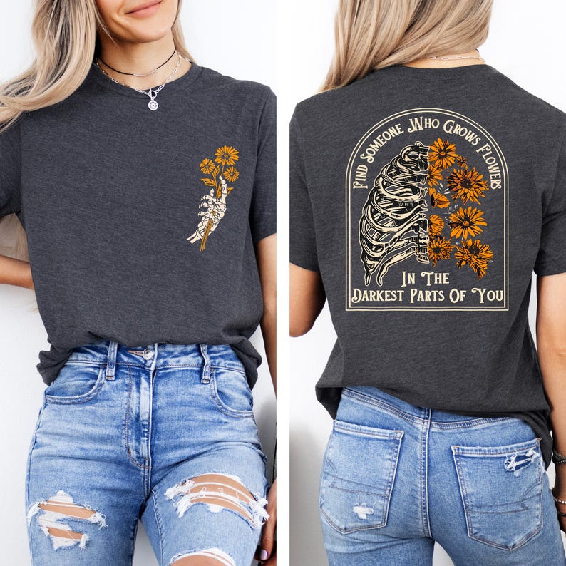 May include: Dark gray t-shirt with a graphic of a skeleton hand holding yellow flowers on the front. The back of the shirt features a design with a rib cage, flowers, and the text "Find Someone Who Grows Flowers In The Darkest Parts Of You."