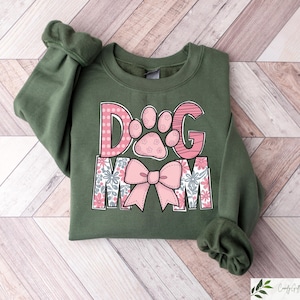 May include: An olive green sweatshirt with the words "DOG MOM" in pink and white. The "D" and "G" are pink with a paw print design. The "O" has a paw print design. The "MOM" has a floral design and a pink bow.