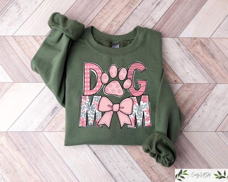 May include: An olive green sweatshirt with the words "DOG MOM" in pink and white. The "D" and "G" are pink with a paw print design. The "O" has a paw print design. The "MOM" has a floral design and a pink bow.