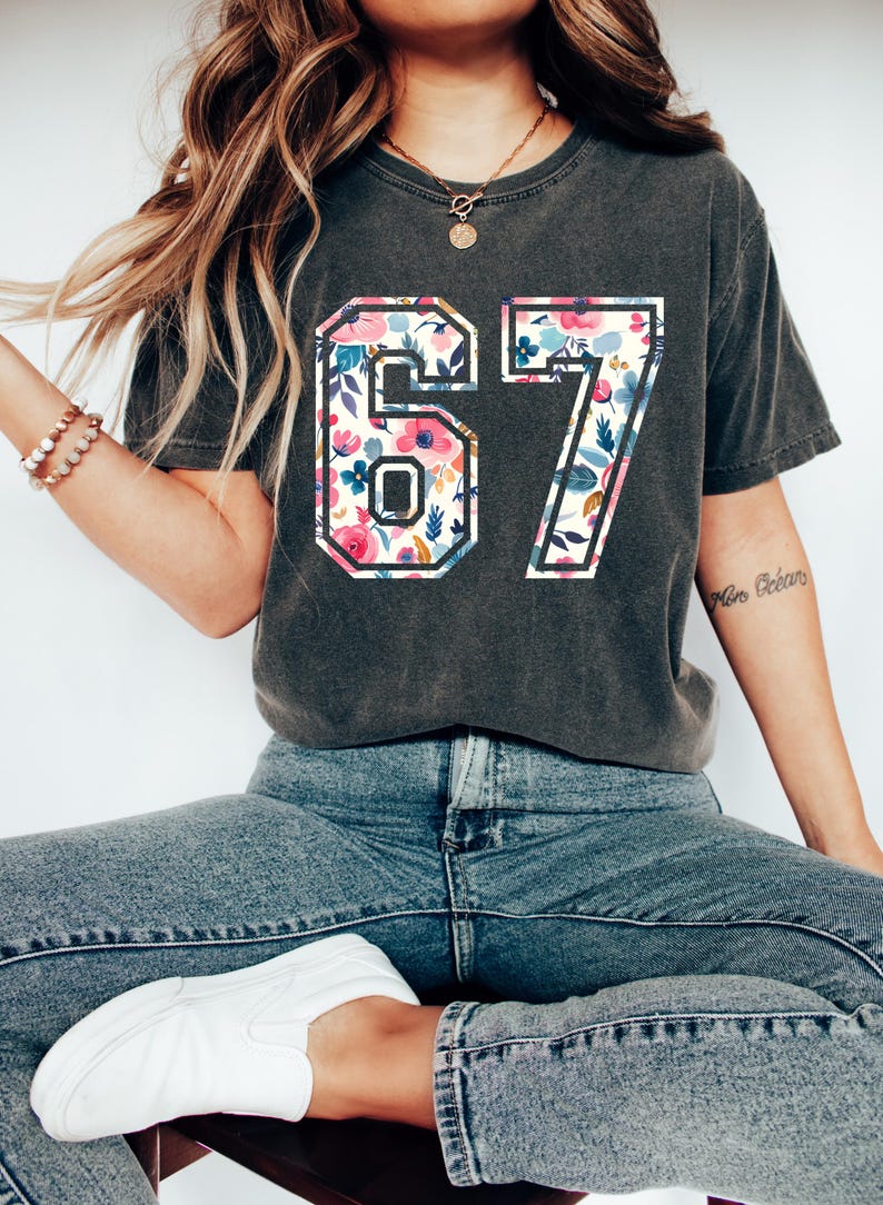 May include: Dark gray t-shirt featuring the number "67" in a floral design with pink, blue, and white flowers. The shirt is paired with blue jeans and white slip-on shoes. The overall style is casual and relaxed.