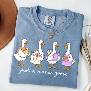 May include: A blue Comfort Colors t-shirt featuring a graphic of four cartoon geese, each with a different accessory. The text "just a mama goose" is printed below the geese. A gold necklace is draped across the shirt.