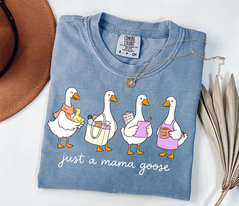 May include: A blue Comfort Colors t-shirt featuring a graphic of four cartoon geese, each with a different accessory. The text "just a mama goose" is printed below the geese. A gold necklace is draped across the shirt.