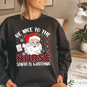 May include: Black sweatshirt with a red and white plaid design. The text "Be Nice To The Nurse Santa Is Watching" is printed on the front of the sweatshirt. A cartoon image of Santa Claus is above the text.