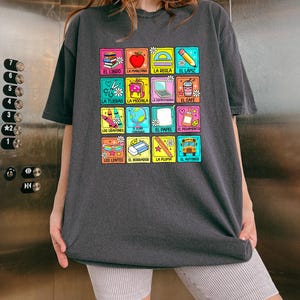 May include: Dark grey t-shirt with a grid of colourful squares, each displaying Spanish words and school-related illustrations. Images include a book, apple, scissors, and a school bus. The text "Comfy Gift Shop" is at the bottom of the image.