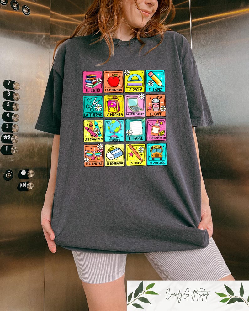May include: Dark grey t-shirt with a grid of colourful squares, each displaying Spanish words and school-related illustrations. Images include a book, apple, scissors, and a school bus. The text "Comfy Gift Shop" is at the bottom of the image.