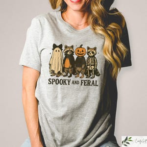 May include: A light grey t-shirt with a Halloween graphic. The design shows a ghost, a raccoon, a pumpkin-headed raccoon, and a skeleton raccoon. The words "Spooky and Feral" are printed beneath the characters. The t-shirt is a casual wear item.