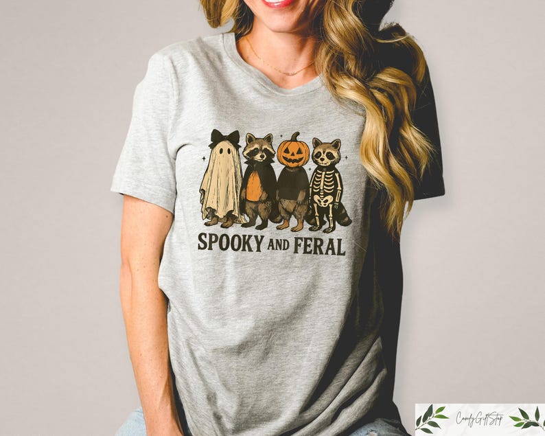 May include: A light grey t-shirt with a Halloween graphic. The design shows a ghost, a raccoon, a pumpkin-headed raccoon, and a skeleton raccoon. The words "Spooky and Feral" are printed beneath the characters. The t-shirt is a casual wear item.
