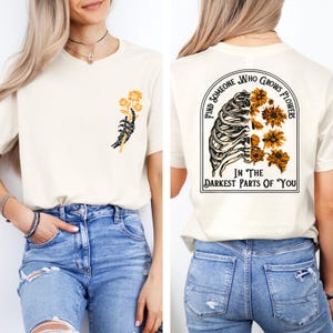 May include: Cream-colored t-shirt with a graphic on the front of a skeleton hand holding yellow flowers. The back of the shirt features a rib cage design with yellow flowers and the text "Find Someone Who Grows Flowers In The Darkest Parts Of You."