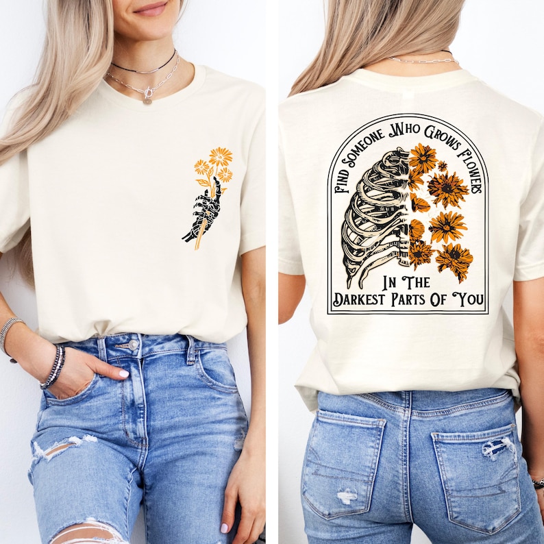 May include: Cream-colored t-shirt with a graphic on the front of a skeleton hand holding yellow flowers. The back of the shirt features a rib cage design with yellow flowers and the text "Find Someone Who Grows Flowers In The Darkest Parts Of You."