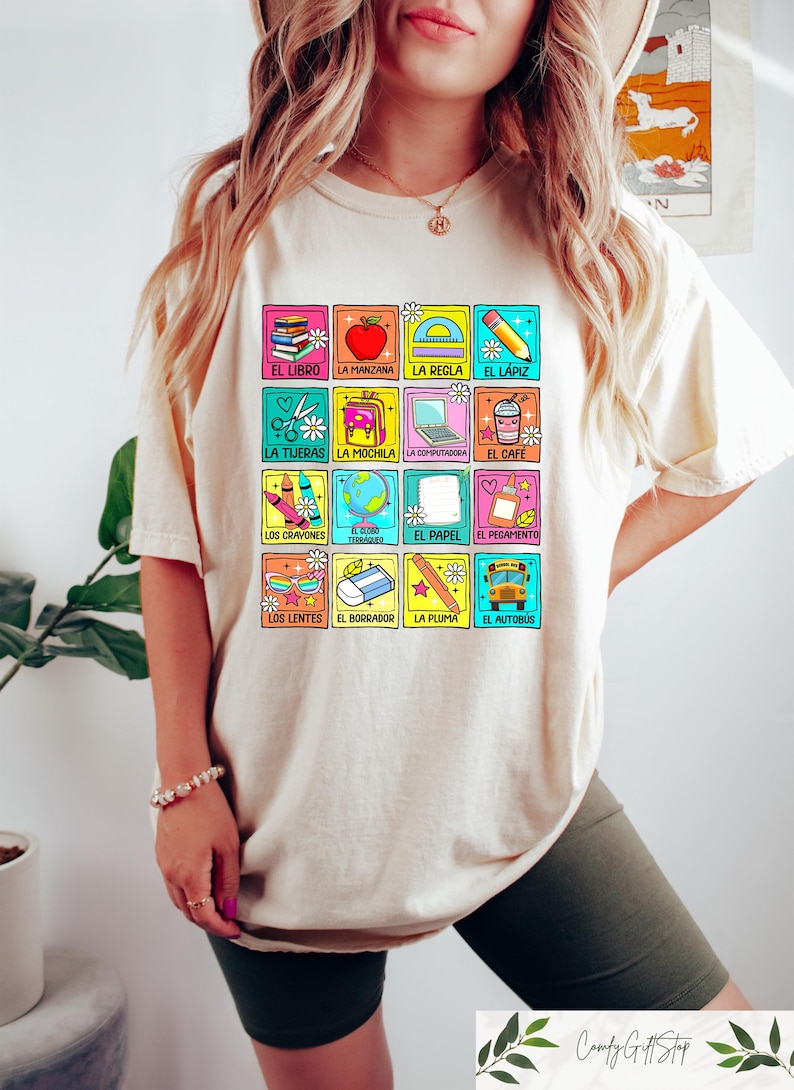 May include: Beige t-shirt with a grid of colourful school-themed images and Spanish words. The images include a book, apple, ruler, pencil, scissors, backpack, laptop, coffee, crayons, globe, paper, glue, glasses, eraser, pen, and school bus.
