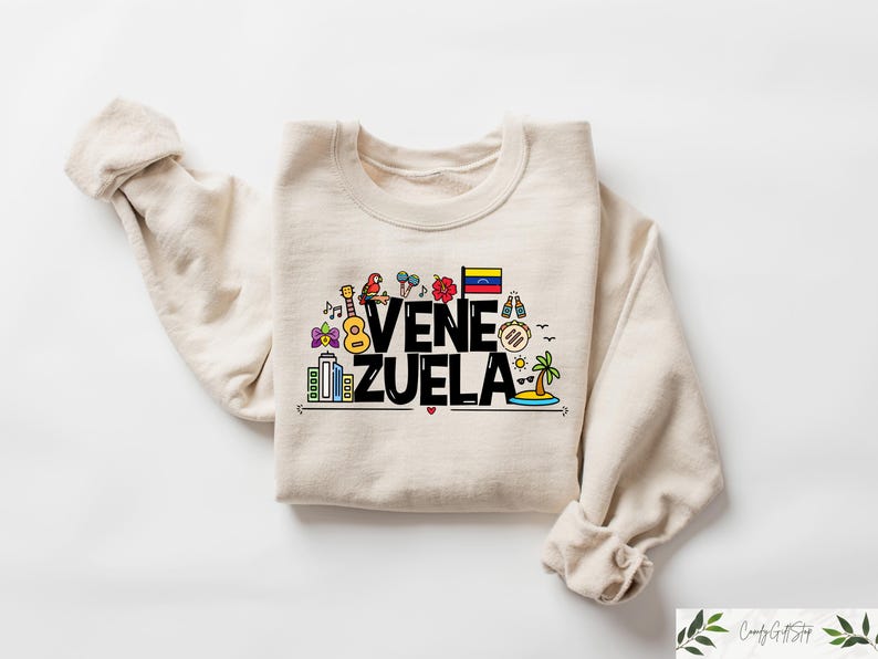 May include: A beige sweatshirt with a black graphic design that says "VENEZUELA" with colorful illustrations of Venezuelan cultural icons, including a guitar, a flag, and a arepa.
