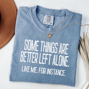 May include: A blue t-shirt with the text "SOME THINGS ARE BETTER LEFT ALONE. LIKE ME, FOR INSTANCE." in white. A gold necklace with a pendant is draped across the shirt. A brown hat is visible in the background.