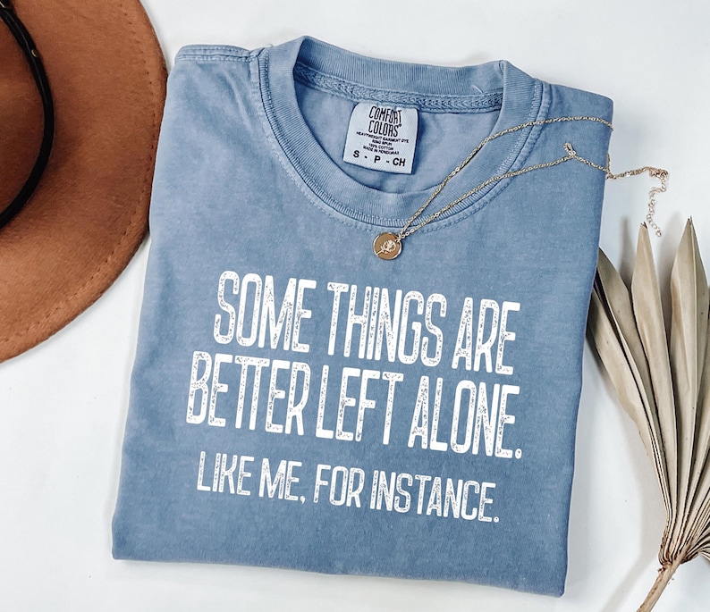 May include: A blue t-shirt with the text "SOME THINGS ARE BETTER LEFT ALONE. LIKE ME, FOR INSTANCE." in white. A gold necklace with a pendant is draped across the shirt. A brown hat is visible in the background.