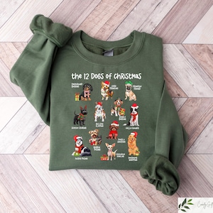 May include: An olive green sweatshirt featuring the text "the 12 DOGS of CHRISTMAS" above illustrations of various dog breeds wearing Santa hats. The sweatshirt is made of a soft material and has ribbed cuffs and a crew neck.