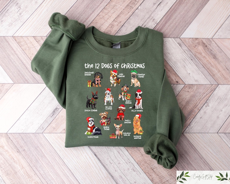 May include: An olive green sweatshirt featuring the text "the 12 DOGS of CHRISTMAS" above illustrations of various dog breeds wearing Santa hats. The sweatshirt is made of a soft material and has ribbed cuffs and a crew neck.