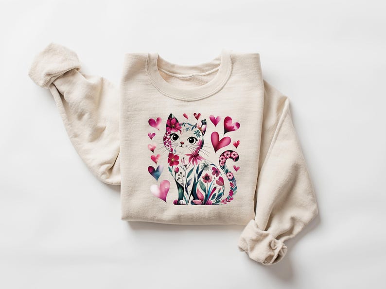 May include: A cream-colored sweatshirt featuring a watercolor illustration of a cat adorned with flowers and surrounded by pink and blue hearts. The cat's design includes floral patterns and heart accents, creating a whimsical and romantic theme.