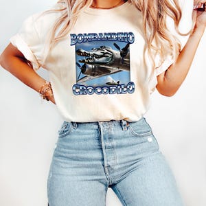 Puede incluir: Camiseta color crema con un dise&ntilde;o gr&aacute;fico de un avi&oacute;n con cabeza de cocodrilo y el texto "BOMBANDINO CROCODILO" en azul. La camiseta se combina con vaqueros azul claro.