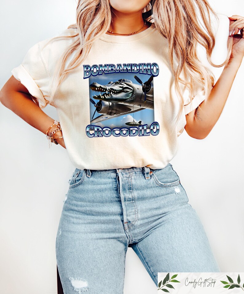 Puede incluir: Camiseta color crema con un dise&ntilde;o gr&aacute;fico de un avi&oacute;n con cabeza de cocodrilo y el texto "BOMBANDINO CROCODILO" en azul. La camiseta se combina con vaqueros azul claro.