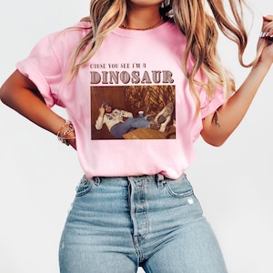 May include: A pink t-shirt with the text "Cause you see I'm a Dinosaur" in brown lettering. The shirt features a vintage photo of a man wearing a cowboy hat and a plaid shirt.