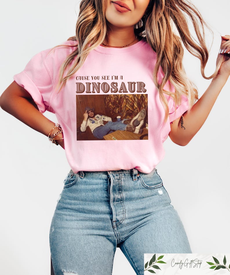 May include: A pink t-shirt with the text "Cause you see I'm a Dinosaur" in brown lettering. The shirt features a vintage photo of a man wearing a cowboy hat and a plaid shirt.