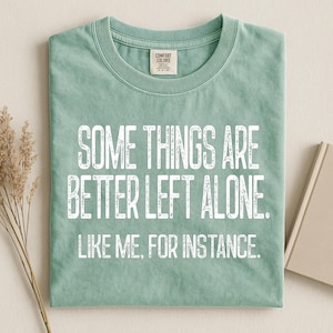 May include: A sage green t-shirt with the text "SOME THINGS ARE BETTER LEFT ALONE. LIKE ME, FOR INSTANCE." in white, centered on the front. The shirt is folded neatly, showcasing the crew neck and the Comfort Colors brand tag.