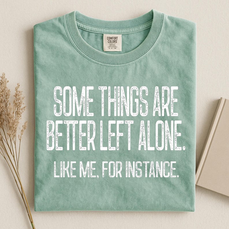 May include: A sage green t-shirt with the text "SOME THINGS ARE BETTER LEFT ALONE. LIKE ME, FOR INSTANCE." in white, centered on the front. The shirt is folded neatly, showcasing the crew neck and the Comfort Colors brand tag.