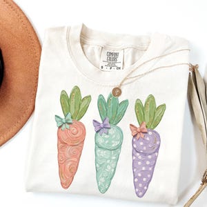 May include: Cream-colored t-shirt featuring three illustrated carrots in pastel colors. Each carrot has green leafy tops and a small bow. A gold necklace with a pendant rests on the shirt. The shirt has a Comfort Colors tag.