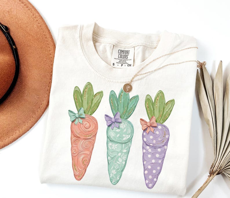 May include: Cream-colored t-shirt featuring three illustrated carrots in pastel colors. Each carrot has green leafy tops and a small bow. A gold necklace with a pendant rests on the shirt. The shirt has a Comfort Colors tag.