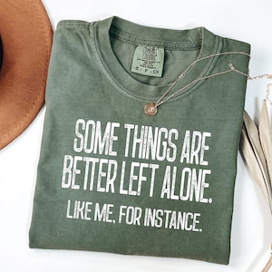 Puede incluir: Camiseta verde oliva con el texto "SOME THINGS ARE BETTER LEFT ALONE. LIKE ME, FOR INSTANCE." Un collar dorado con un colgante descansa sobre la camiseta. Un sombrero marrón y una hoja de palma seca están en el fondo.