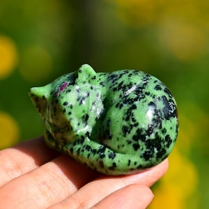 May include: A green and black stone cat figurine in a curled-up sleeping position. The cat has a polished finish and small red accents. The figurine is held in a hand against a blurred green and yellow background.