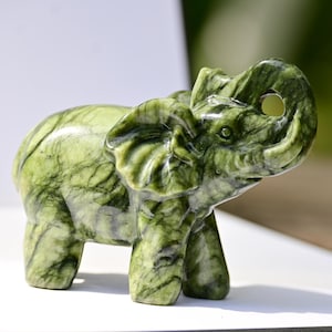 Könnte beinhalten: Kleine, geschnitzte Elefantenfigur in Grüntönen. Der Elefant aus Stein hat einen erhobenen Rüssel und detaillierte Merkmale. Die Figur steht auf einer weißen Oberfläche mit verschwommenem Hintergrund.
