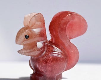 May include: A small, carved squirrel figurine made from a translucent, reddish-pink stone. The sculpture features a smooth, polished surface, a prominent bushy tail, pointed ears, and a small black dot representing the eye on its side profile.