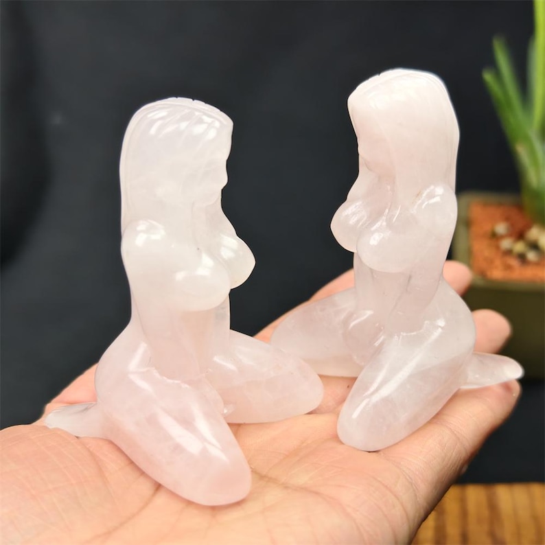 May include: Two translucent, light pink crystal mermaid figurines. The mermaids are kneeling with their hands on their knees. The figurines are held in a person's hand against a dark background.
