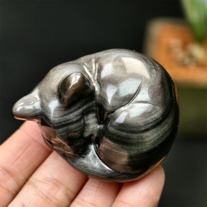 May include: A polished, silver sheen obsidian sculpture of a sleeping animal, possibly a bear or a cat. The stone has a swirling pattern and is held in a hand. The sculpture is smooth and rounded.