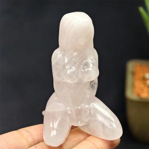 May include: A carved rose quartz figurine depicting a kneeling figure with long hair. The sculpture is translucent with a soft pink hue, and the details of the form are visible. The figurine is held in a hand against a dark background.