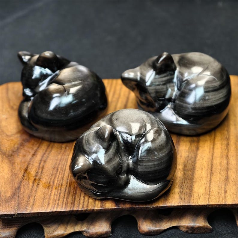 May include: Three polished black obsidian cat figurines in a curled sleeping position. The cats have a reflective, striped pattern and sit on a wooden surface. The background is dark.