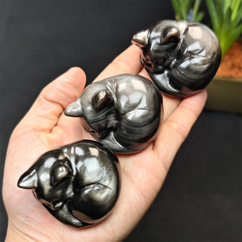 May include: Three polished obsidian cat figurines, each curled up in a sleeping position. The cats are black with silver streaks, and are held in a hand. The figurines are smooth and reflective, with detailed features.