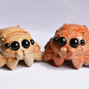 May include: Two carved stone spider figurines with smooth, polished surfaces. The left spider is light beige with tan striations, while the right spider is a deep reddish-orange. Both feature four prominent, glossy black circular eyes.
