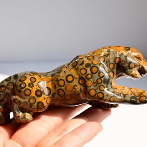 May include: A polished stone figurine carved into the shape of a leaping leopard. The surface features a natural pattern of circular spots in shades of mustard yellow, dark brown, and olive green, creating a distinct spotted appearance.