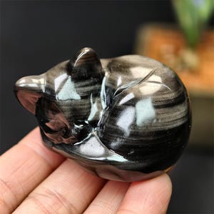 May include: A polished, black obsidian cat figurine in a curled sleeping position. The stone has a swirling pattern of black and gray, with a glossy finish. The cat's ear and head are clearly defined, showcasing the craftsmanship of the sculpture.