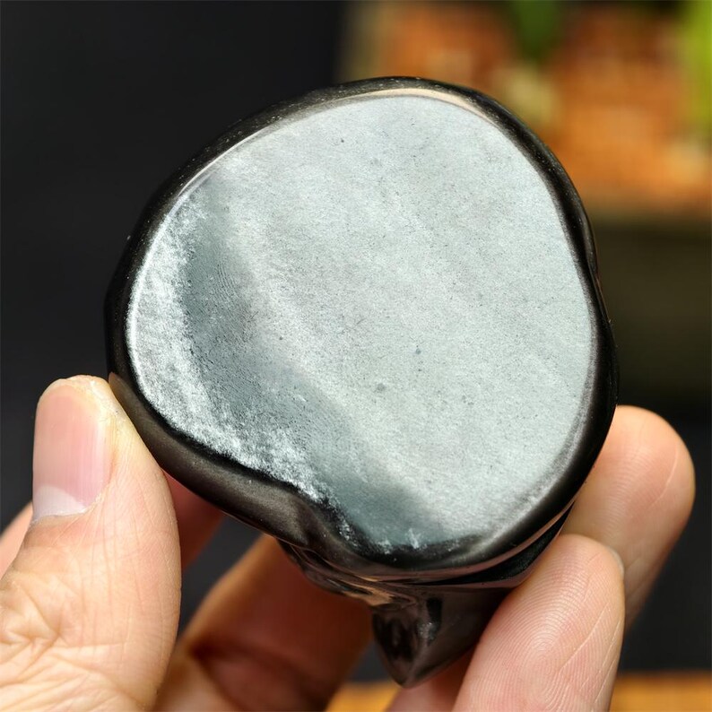 May include: A polished, black obsidian skull carving. The skull has a smooth, rounded top with a reflective, silvery sheen. The carving is held in a hand, showcasing the detailed craftsmanship and the natural variations in the stone.