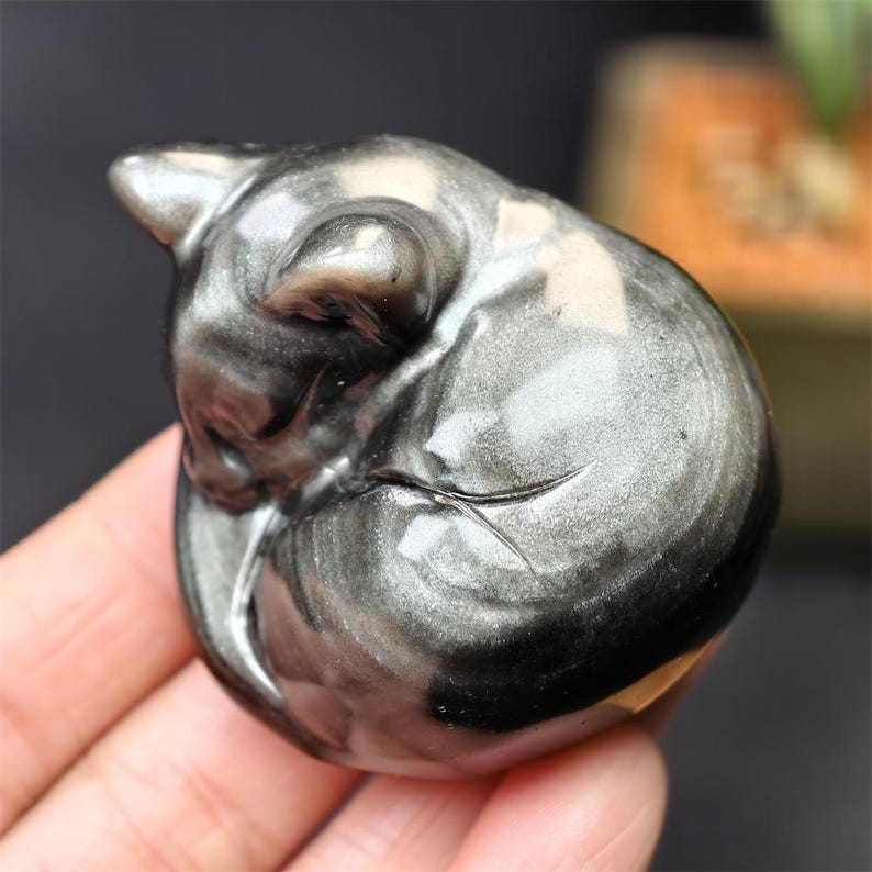 May include: A polished, silver-colored gemstone carved into the shape of a sleeping cat. The cat is curled up, with its head resting on its paws. The stone has a metallic sheen and a smooth, rounded form.