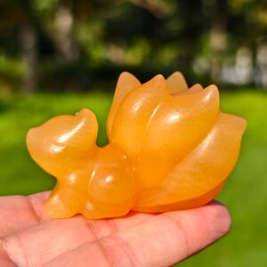 May include: A carved orange crystal fox figurine. The fox is depicted in a lying down position with a large, fanned tail. The figurine is smooth and polished, with a translucent quality. The background is blurred green.