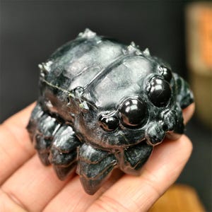 May include: A dark green, carved stone spider figurine with multiple eyes and legs. The spider is detailed with a textured body and is held in a hand. The figurine is a decorative item.