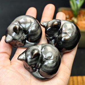 May include: Three polished obsidian cat figurines, each curled in a sleeping position. The black stone has silver streaks, creating a reflective, marbled effect. The cats are held in a hand, showcasing their size and detail.